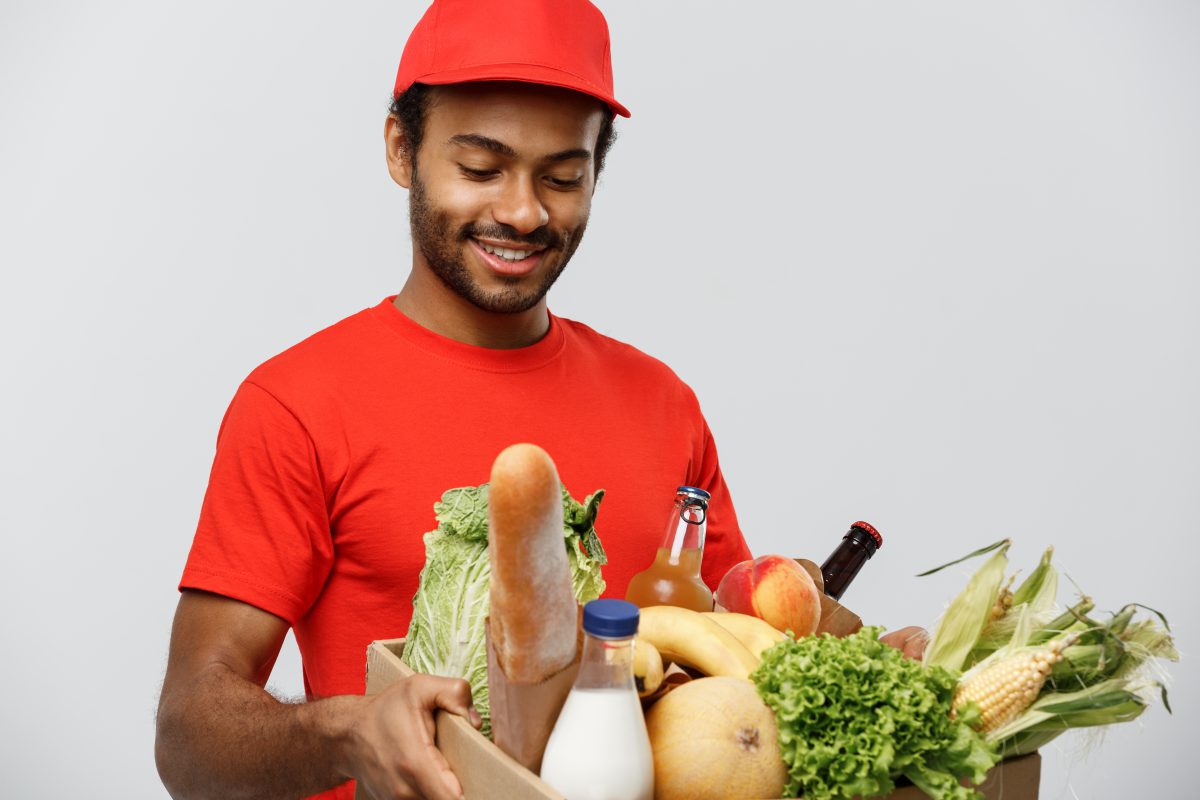Homem com uniforme vermelho segurando caixa com mantimentos e verduras sorrindo
