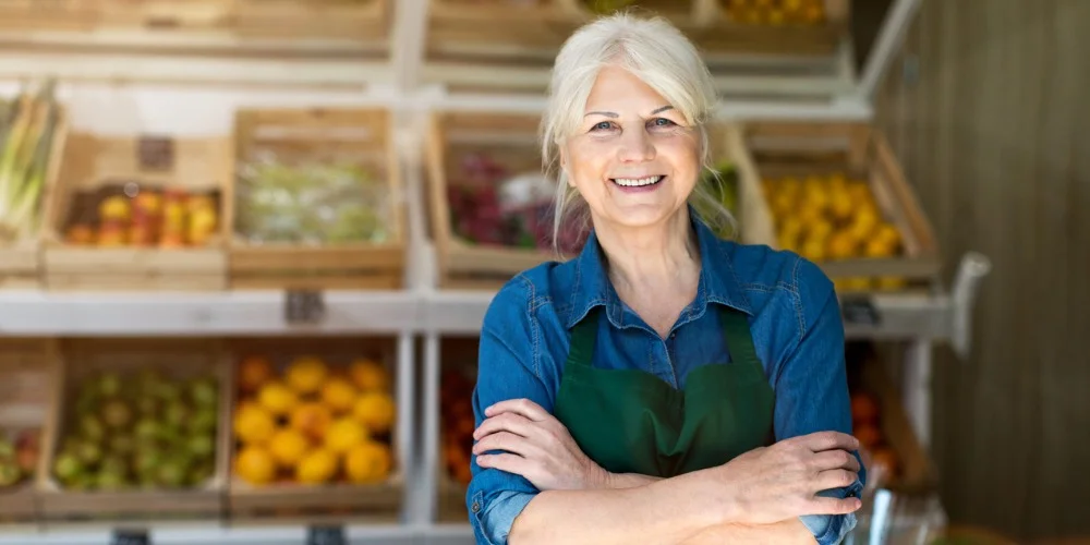 Vendedora de mercado sorrindo com braços cruzados em frente a bancadas de frutas